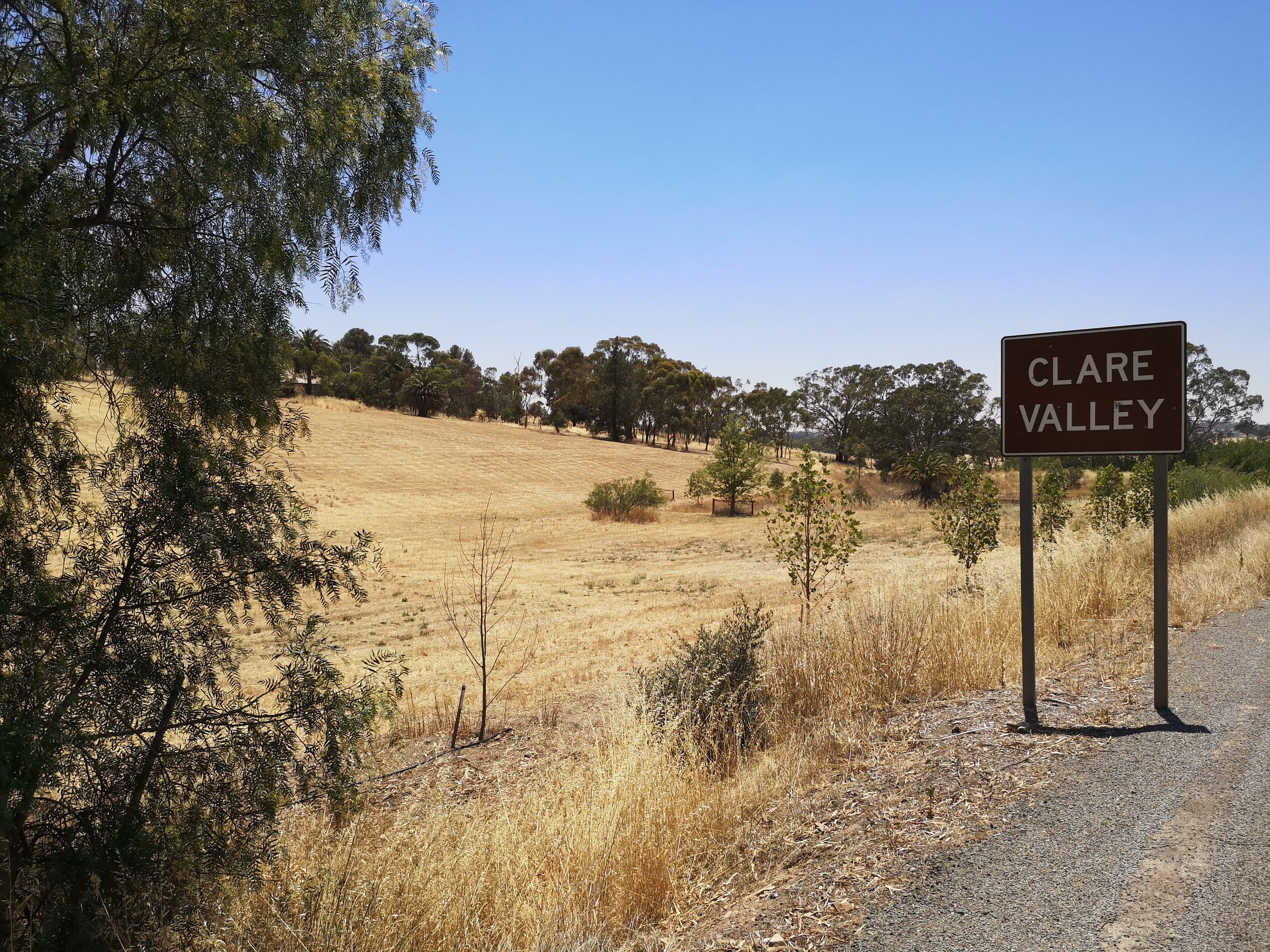Brown sign with white text reading "Clare Valley" on a metal post, with a dry landscape of yellow grass and scattered trees in the background under a clear blue sky. Located just two hours from Adelaide, South Australia.